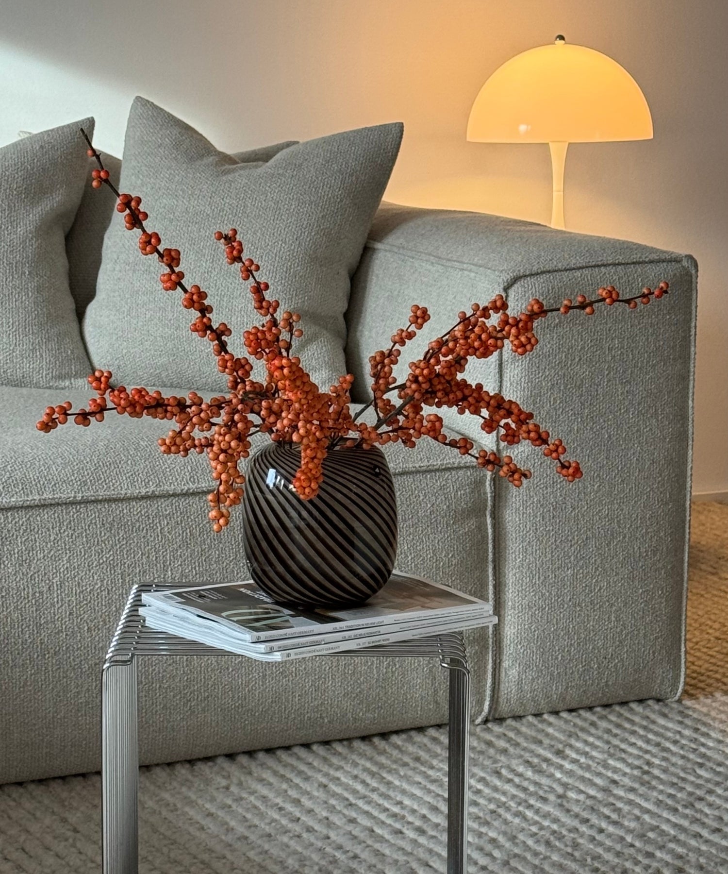 Herbstlich dekoriertes Wohnzimmer mit grauem Sofa, schwarzer Vase mit orangeroten Beeren, Kissen, Zeitschriften und gelber Pilzleuchte