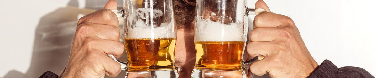 Zwei Männerhände stoßen mit gefüllten Biergläsern an, bernsteinfarbenes Bier, heller Hintergrund mit Schatten