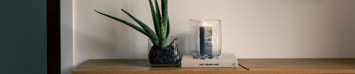 Holztisch mit Sukkulente in Glastopf, Kerze im Glas und Buch mit "EAT WORK SLAY" Schriftzug vor neutraler Wand