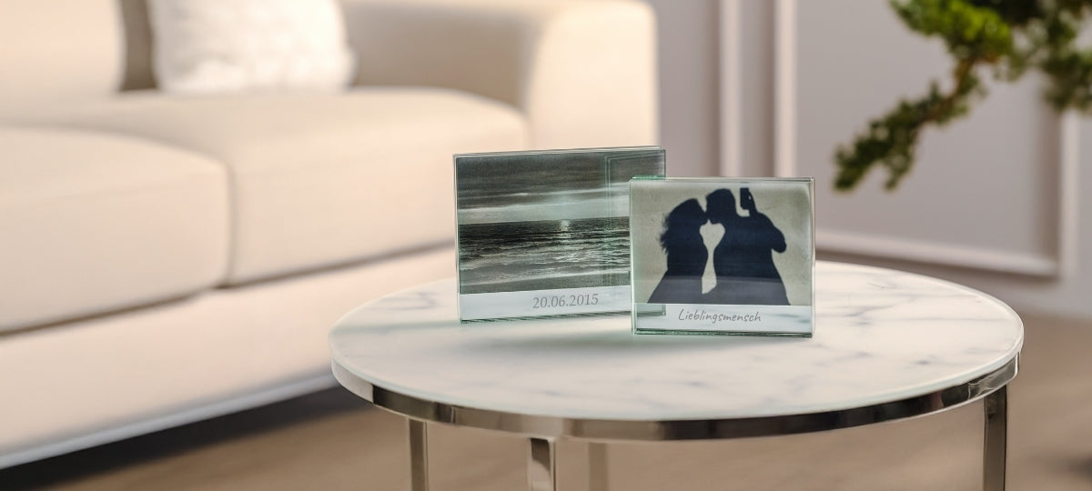 Zwei gravierte Fotorahmen aus Glas auf marmornem Tisch, dahinter helles Wohnzimmer mit Sofa und Pflanze