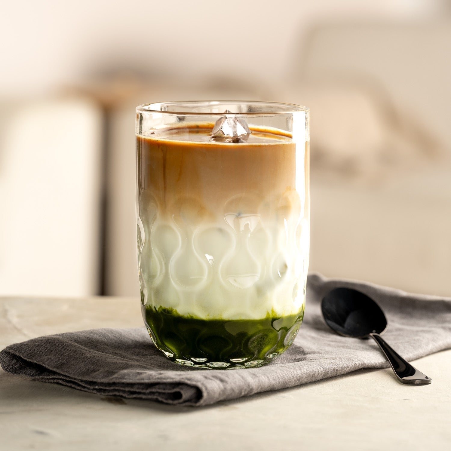 Geschichteter Eiskaffee mit Matcha, Milch und Espresso in einem strukturierten Glas von LEONARDO auf einem grauen Stofftuch mit schwarzem Löffel daneben.