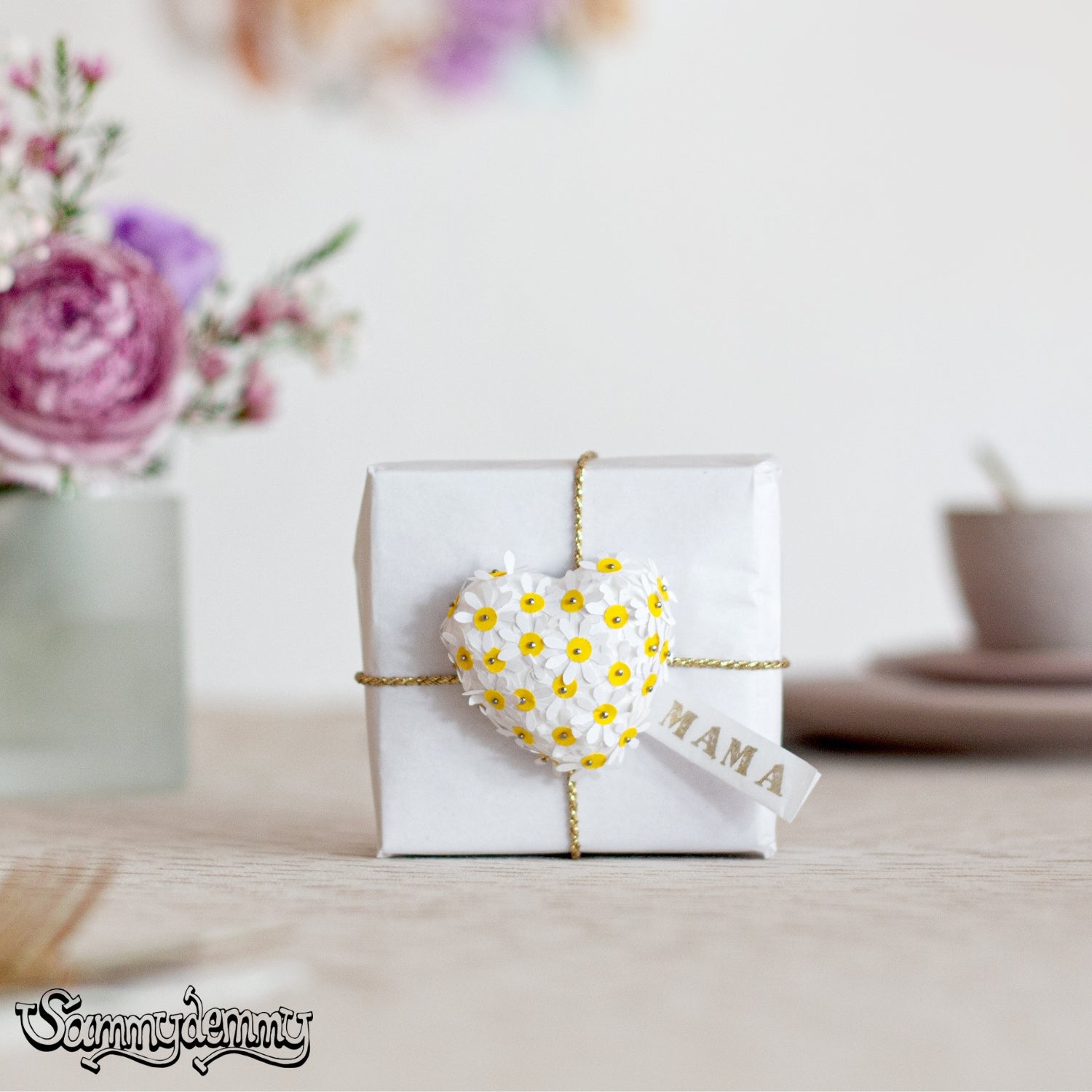 Ein liebevoll verpacktes Geschenk mit weißem Papier, goldener Kordel und einem Herzanhänger aus weißen Blümchen mit gelber Mitte sowie einem „MAMA“-Schild, auf einem gedeckten Tisch mit Blumen und Geschirr im Hintergrund