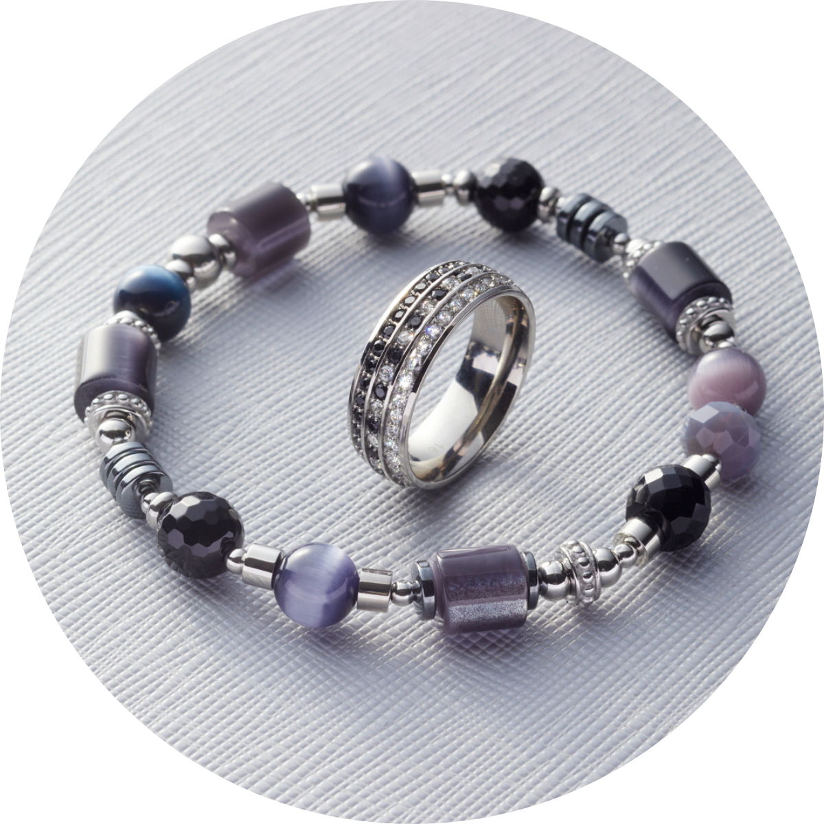 Ein silberfarbenes Armband mit violetten und schwarzen Perlen sowie ein funkelnder Ring liegen auf strukturiertem Stoff.