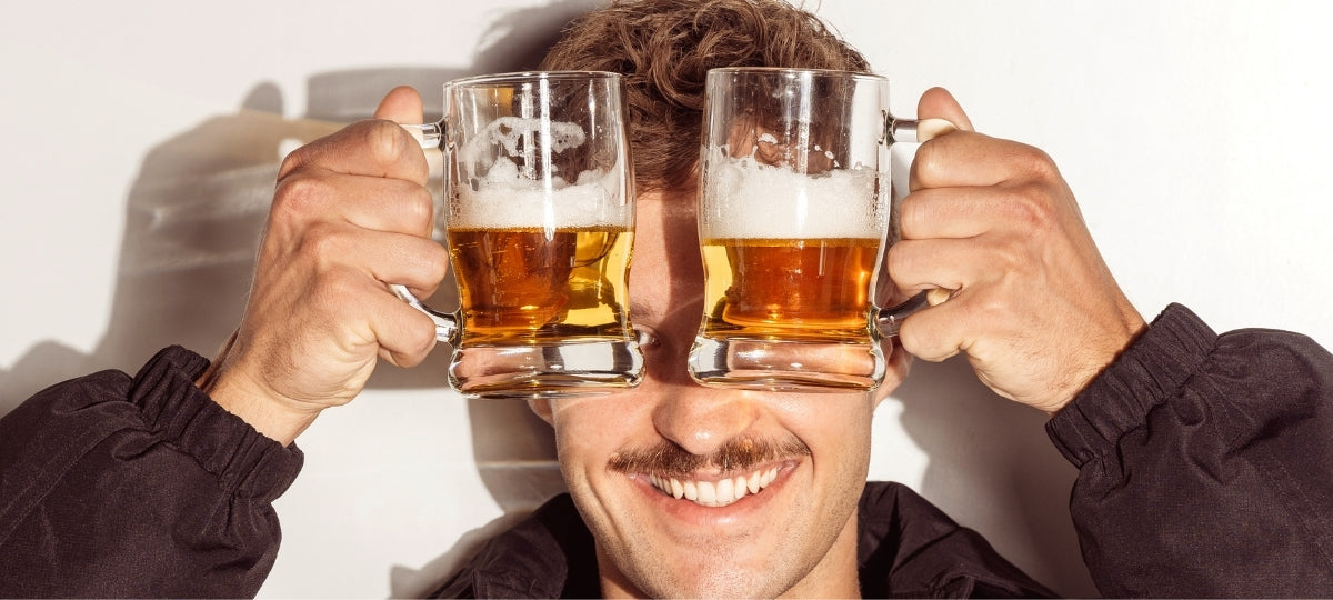 Mann mit Schnurrbart hält zwei volle Biergläser vor sein Gesicht und lächelt breit