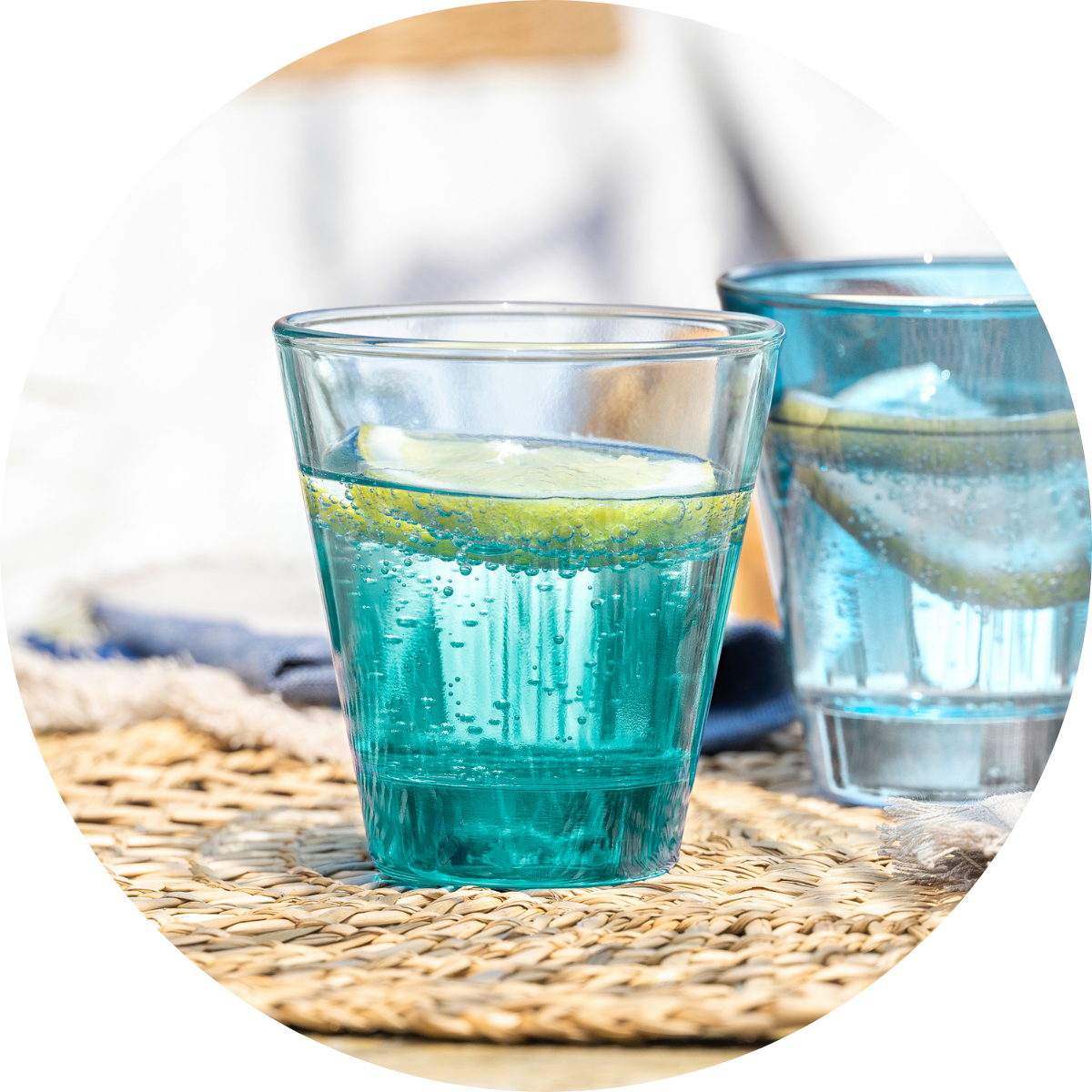 Blaugrünes Wasserglas mit Zitronenscheibe auf geflochtener Unterlage im Sonnenlicht.