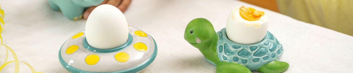 Zwei bunte Kinder-Eierbecher: einer mit Punktmuster und gelben Eiern, einer grüne Schildkröte mit türkisem Halsband