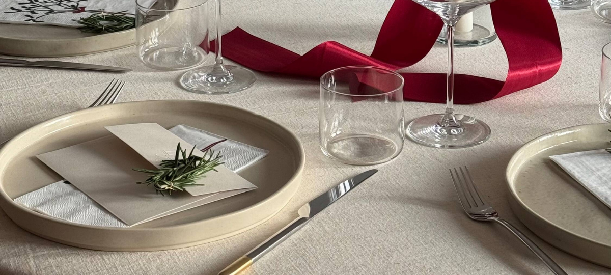Festlich gedeckter Tisch mit beigen Tellern, gefalteten Servietten und einem Rosmarinzweig, daneben Wein- und Wassergläser sowie rotes Geschenkband.