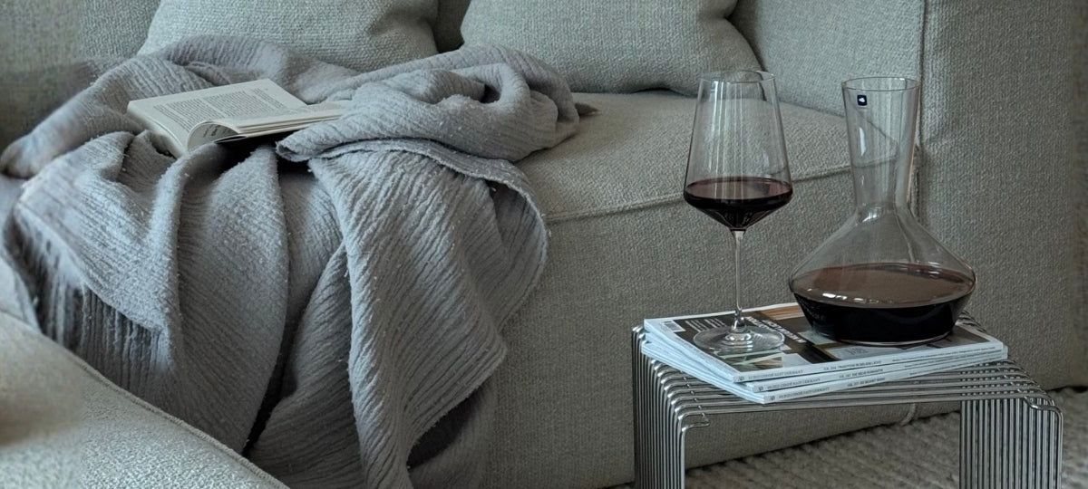 Glaskaraffe mit Rotwein auf Magazinstapel neben Weinglas auf grauem Sofa mit Wolldecke