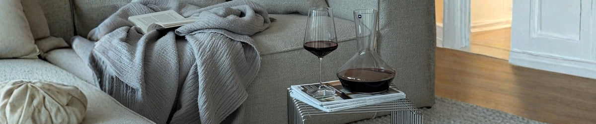 Elegante Karaffen mit Rotwein auf Nachttisch neben grauem Sofa mit Decke, Magazinen und Kerze in warmem Wohnzimmer