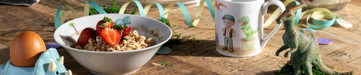 Kinderschale mit Müsli und Erdbeeren, daneben Ei, Becher und grüner Dinosaurier auf Holzuntergrund mit Dekobändern