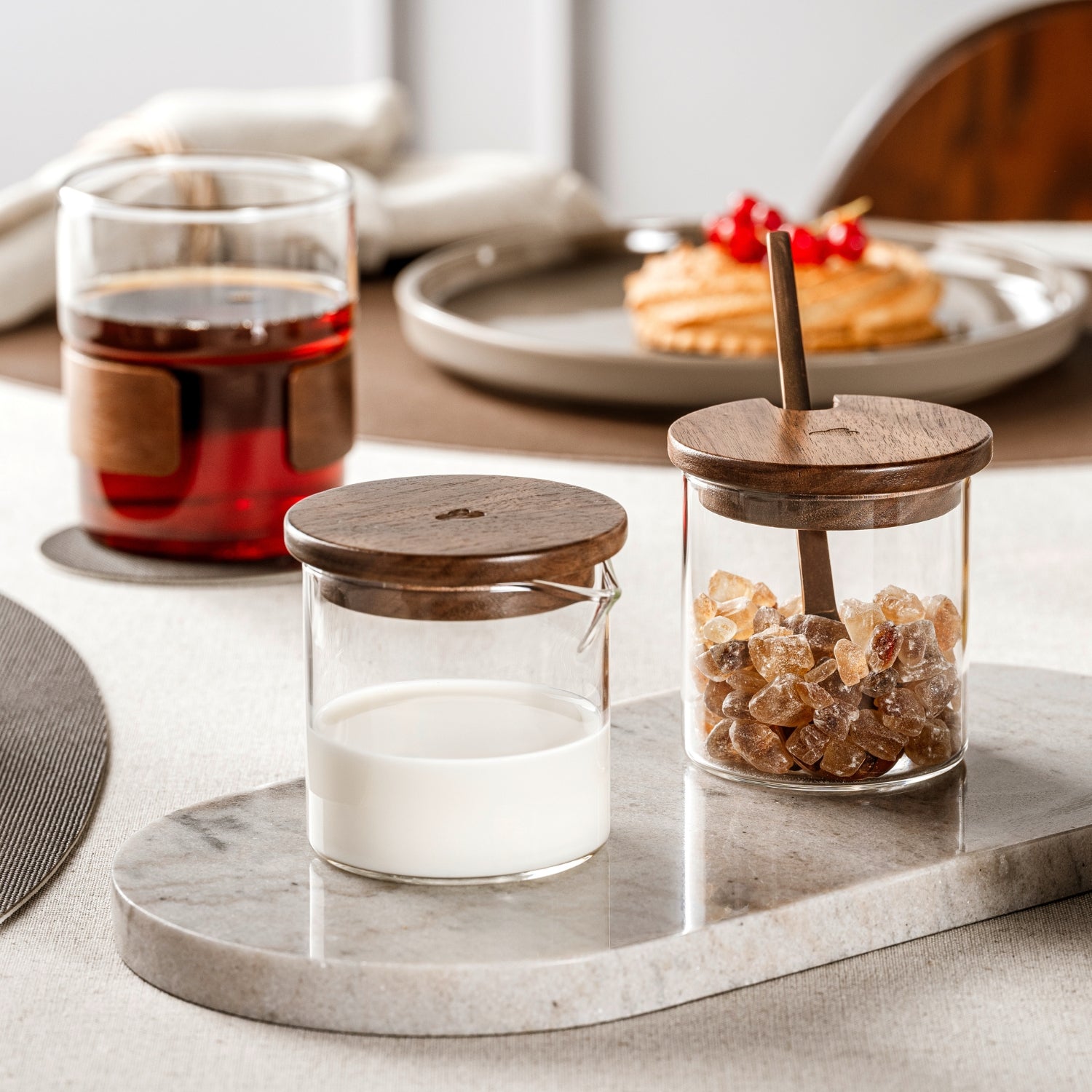Zwei kleine Vorratsgläser aus Glas mit edlen Holzdeckeln stehen auf einer hellen Marmorplatte und sind mit Milch und Kandiszucker gefüllt – perfekt inszeniert für den stilvollen Kaffeegenuss. Im Hintergrund eine gefüllte Teetasse.