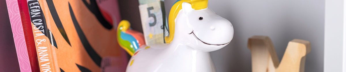Weiße Spardose in Einhornform mit gelbem Horn neben bunten Büchern und Dekoobjekten